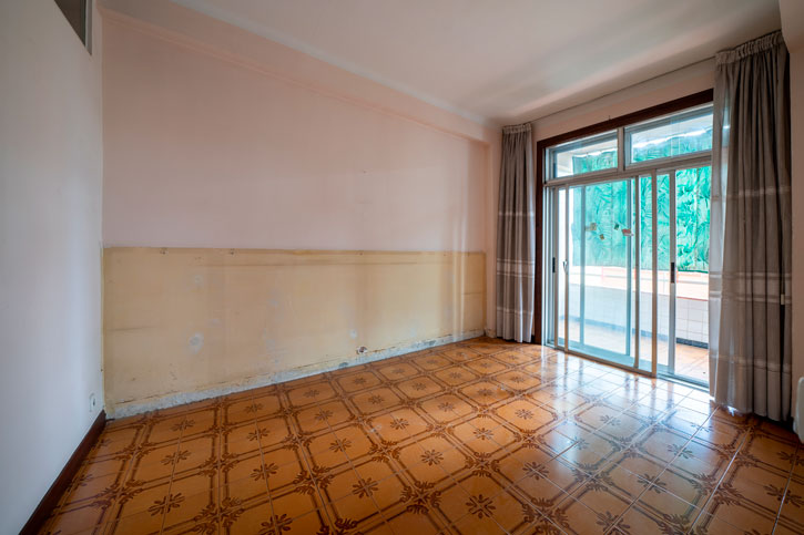 Salón antes de la reforma, con suelo cerámico marrón, carpintería oscura y acceso al balcón.