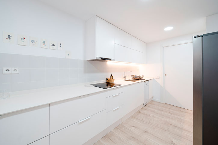 Cocina reformada en blanco con mobiliario lineal, encimera continua y pavimento claro en acabado madera.