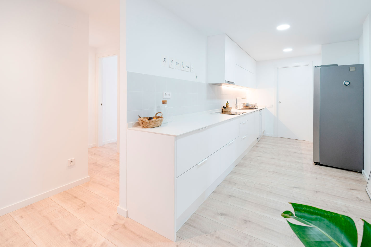 Cocina reformada con mobiliario blanco, encimera lineal, frigorífico gris y pavimento claro.