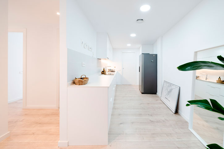 Cocina blanca abierta hacia el salón, con pilar de hormigón visto y pavimento continuo en tono madera.