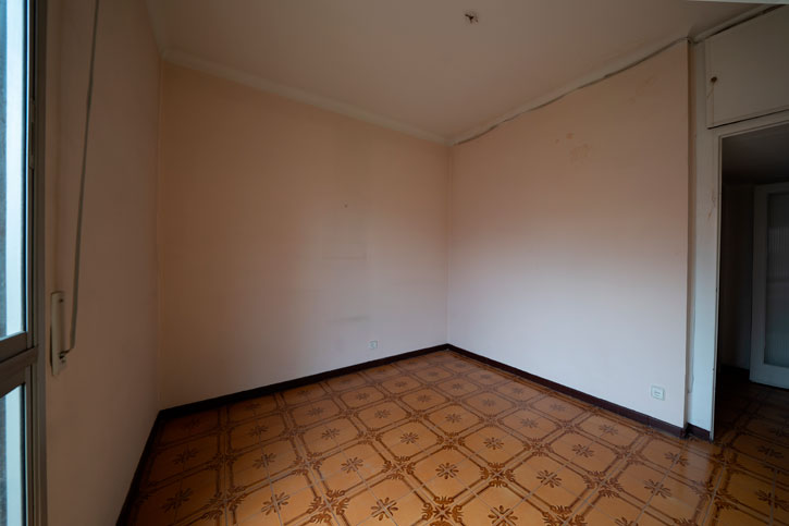Dormitorio antes de la reforma con paredes rosadas, suelo cerámico oscuro y ventana lateral.