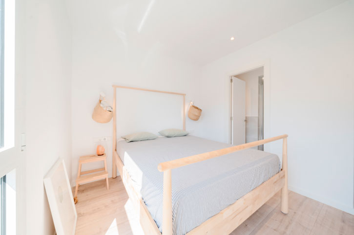 Dormitorio reformado con cama de madera clara, paredes blancas, pavimento en tono madera y luz natural.