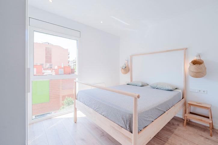 Dormitorio reformado con cama de madera, gran ventana, paredes blancas y pavimento claro.