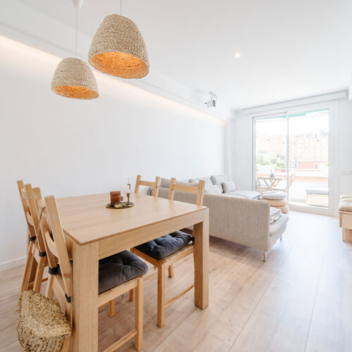 Comedor y salón abiertos con mesa de madera, lámparas de fibras naturales, sofá y salida a la terraza.