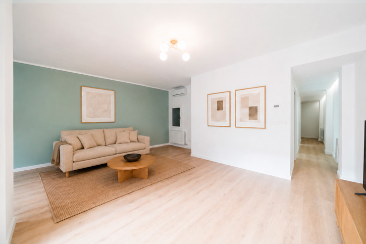 Salón reformado con sofá beige, pared verde y conexión con el pasillo de la vivienda.