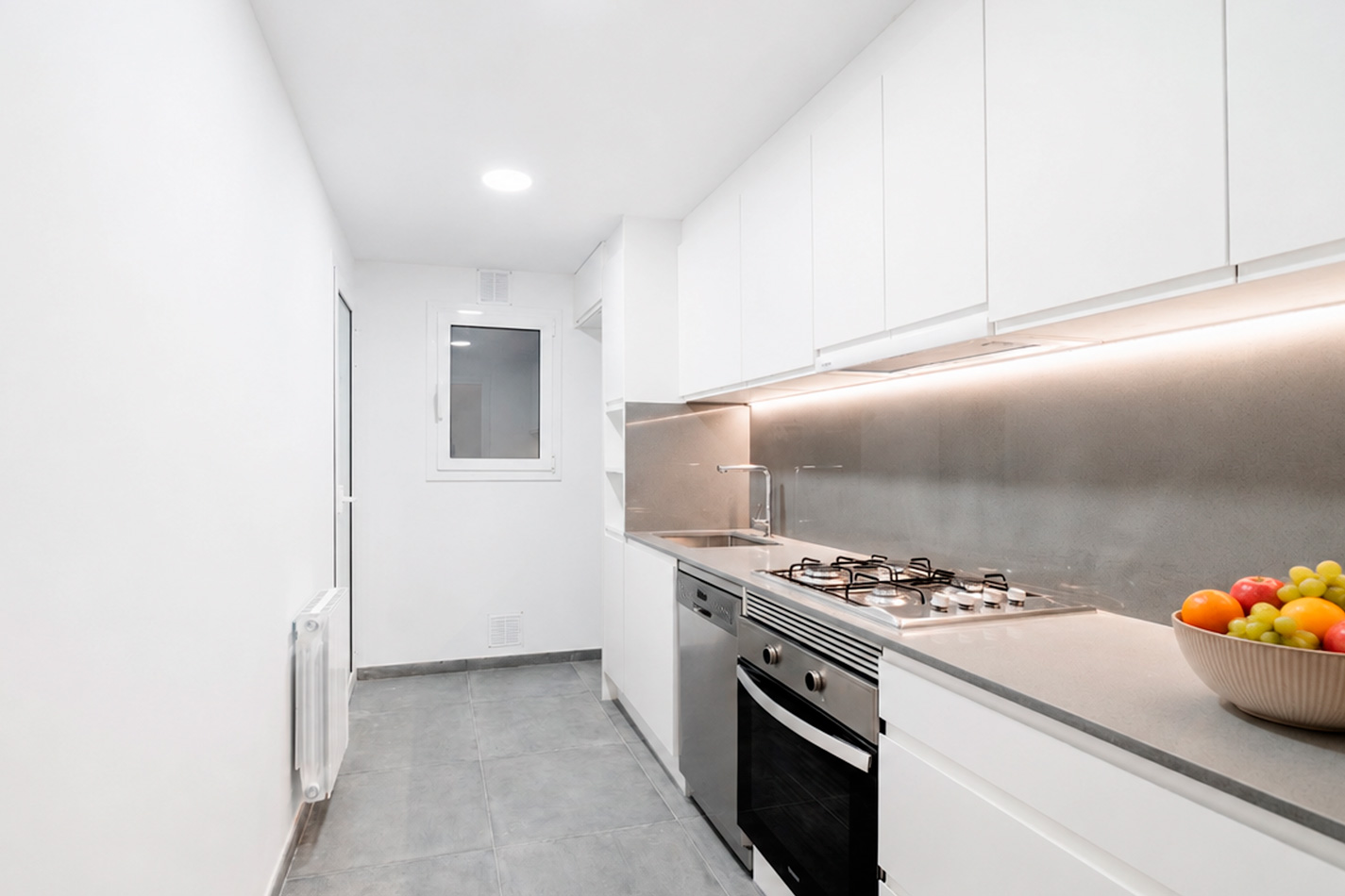 Cocina reformada en el Eixample de Barcelona con muebles blancos, encimera gris, iluminación lineal y pavimento gris